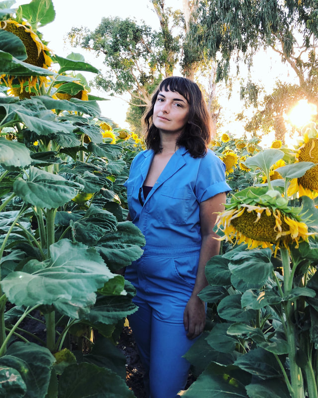 Eva standing in a field of sunflowers with the sun setting off to her right. She has shoulder length brown hair and short bangs. She is wearing a blue mechanics jumpsuit and looking at the camera with a solemn, bemused look and her head tilted to the side. 